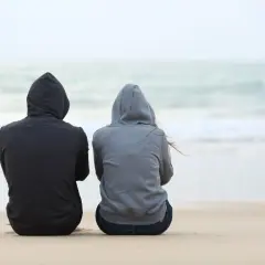 Dwie osoby w bluzach z kapturem siedzą na plaży, patrząc na morze. Czasem spotykamy ludzi, którzy stają się częścią naszych wspomnień.