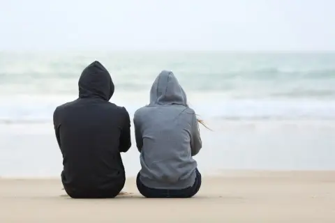 Dwie osoby w bluzach z kapturem siedzą na plaży, patrząc na morze. Czasem spotykamy ludzi, którzy stają się częścią naszych wspomnień.