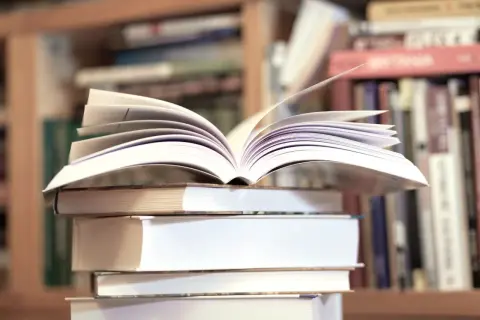 Otwarta książka na stosie innych, w tle regały pełne literatury, w tym klasyka jak "Zbrodnia i kara" gatunek.
