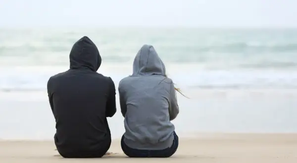 Dwie osoby w bluzach z kapturem siedzą na plaży, patrząc na morze. Czasem spotykamy ludzi, którzy stają się częścią naszych wspomnień.