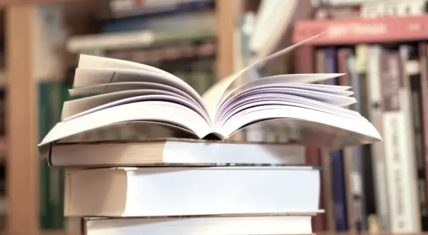 Otwarta książka na stosie innych, w tle regały pełne literatury, w tym klasyka jak "Zbrodnia i kara" gatunek.