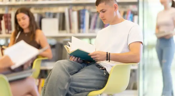 Młody mężczyzna czyta książkę w bibliotece, jakby szukał znaczenia tytułu "Przedwiośnie". W tle inne osoby pochłonięte lekturą.