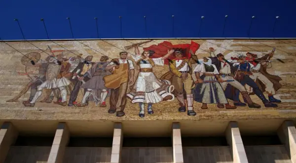 Mozaika w stylu socrealizmu przedstawia grupę ludzi w strojach ludowych, z bronią i flagami, symbolizujących walkę i jedność.