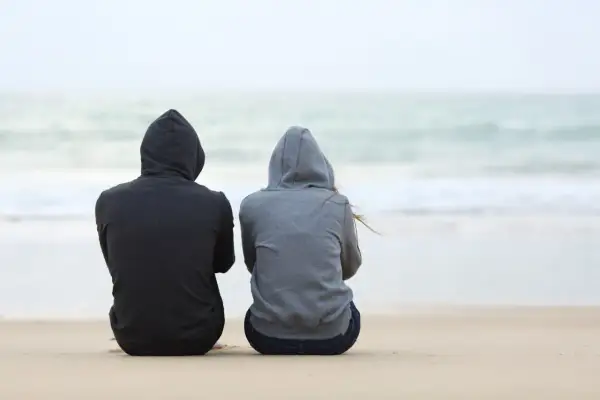 Dwie osoby w bluzach z kapturem siedzą na plaży, patrząc na morze. Czasem spotykamy ludzi, którzy stają się częścią naszych wspomnień.